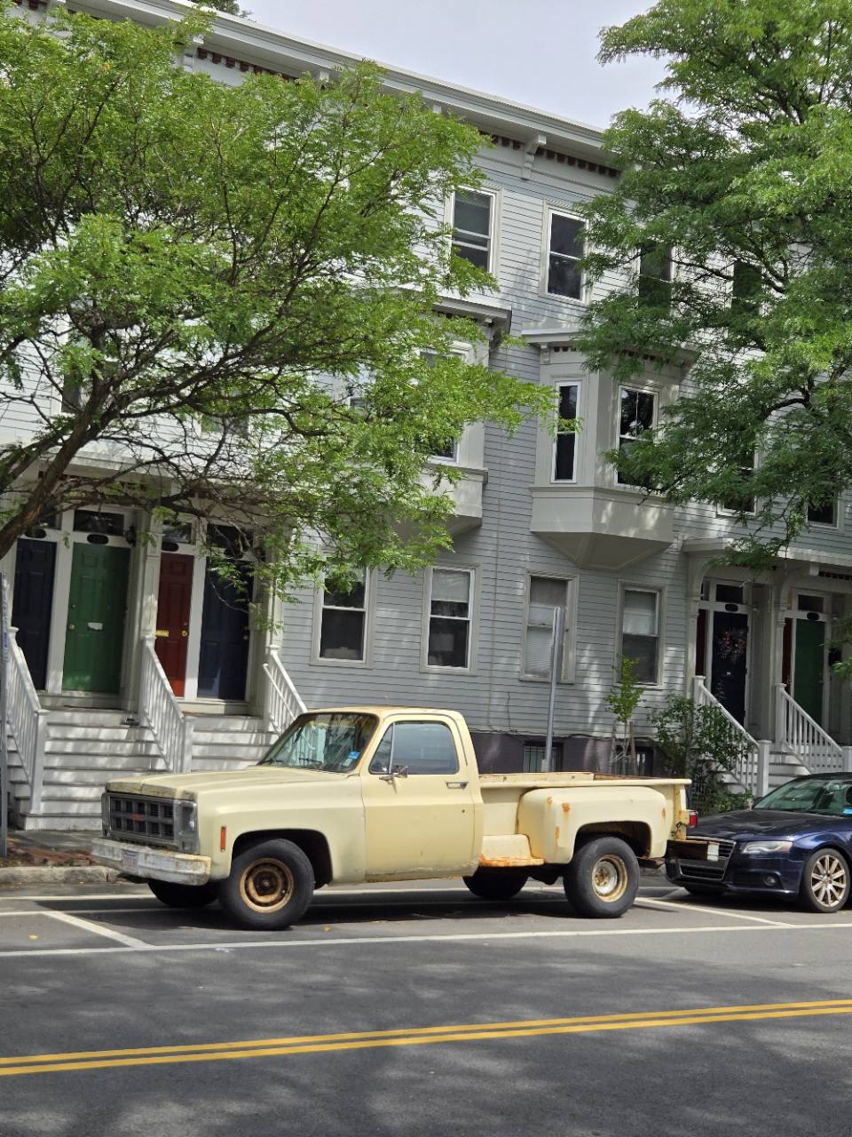 A pale yellow pickup truck parked in front of gray houses and leafy green trees.