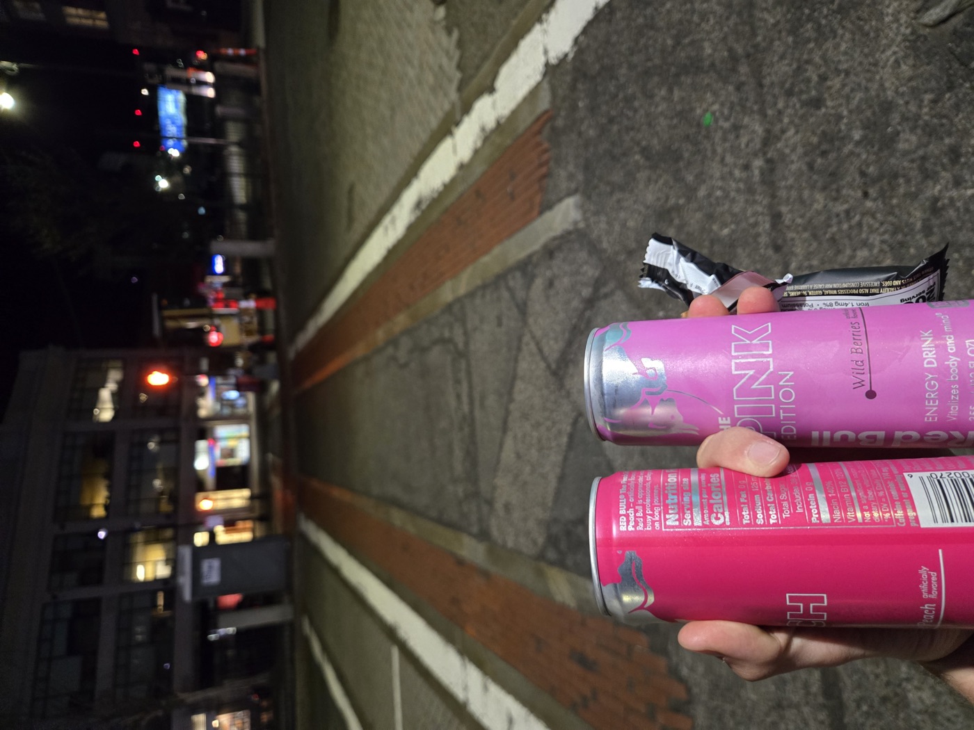Two pink energy drink cans held up at an empty crosswalk at night.