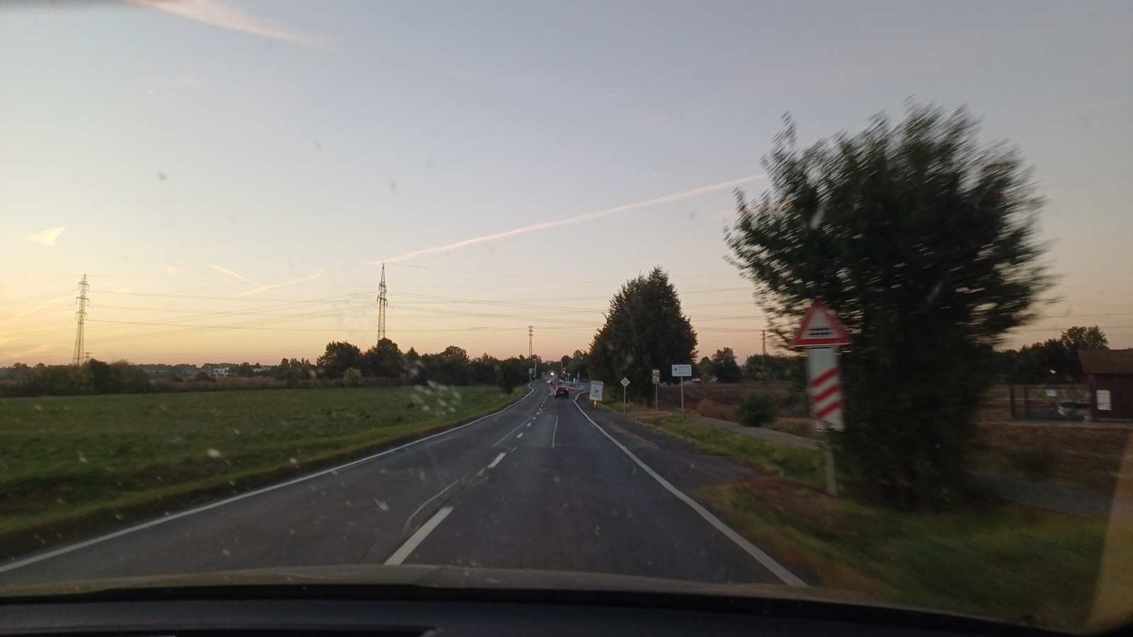 A two-lane country road at dusk, seen through a windshield, with power lines and a railway crossing sign.