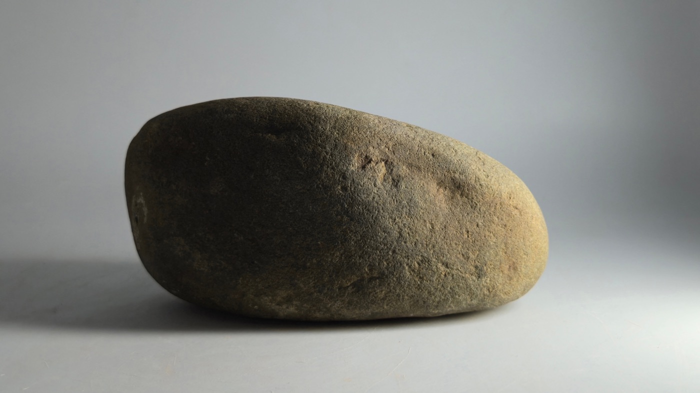 A smooth gray-brown stone resting on a pale surface, lit from the side.